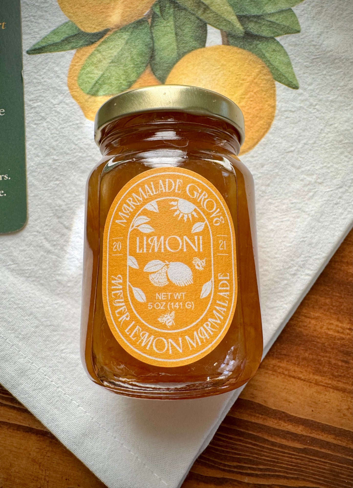 Close up view of a jar of Limoni honey meyer lemon marmalade on top of a lemon branch tea towel on a wood table