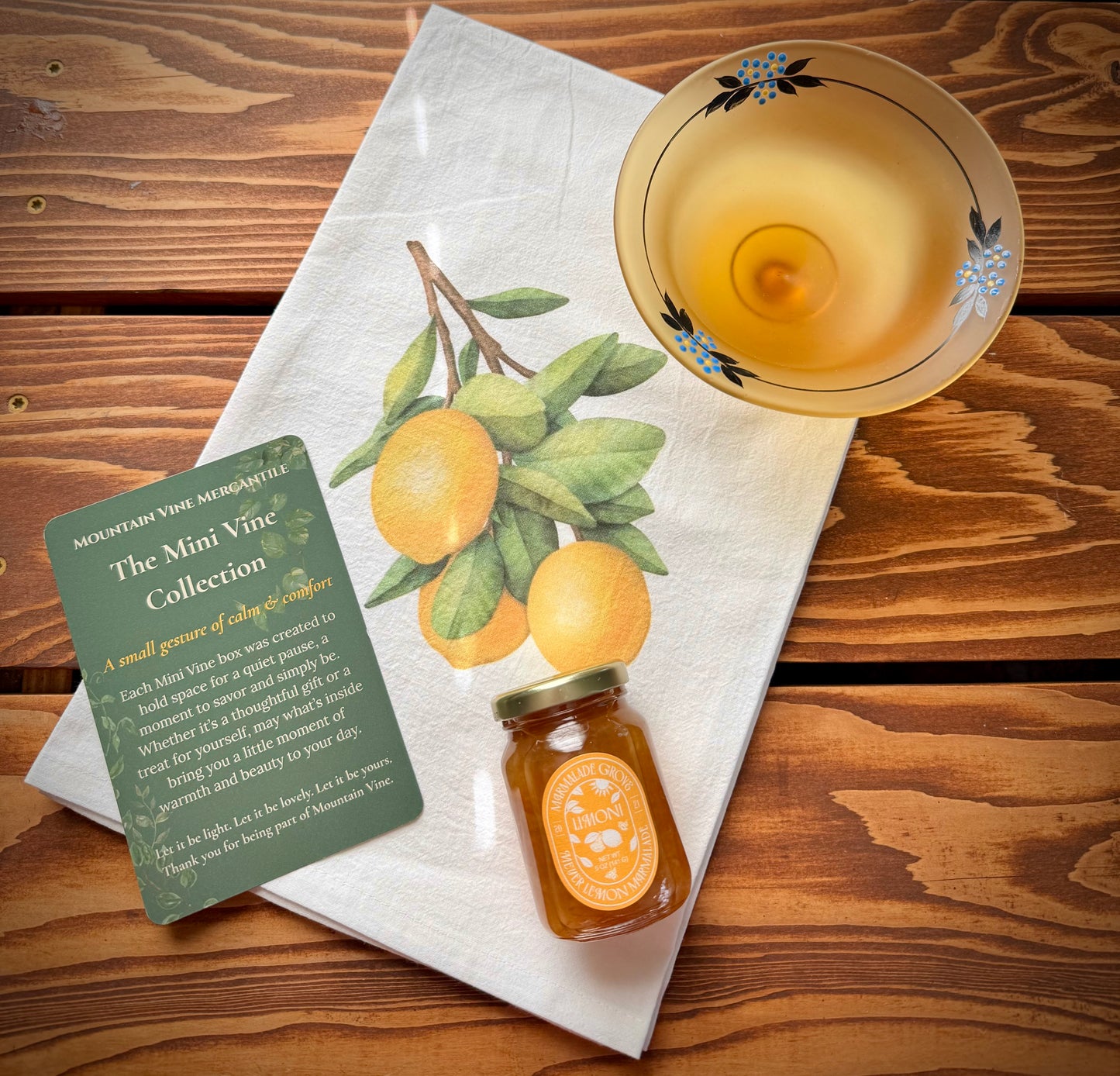 A bright white flour sack tea towel with lemon branch print, a jar of limoni marmalade by Marmalade Grove, a frosted yellow vintage pedestal dish with painted blue flowers and a green product insert card on a wood background