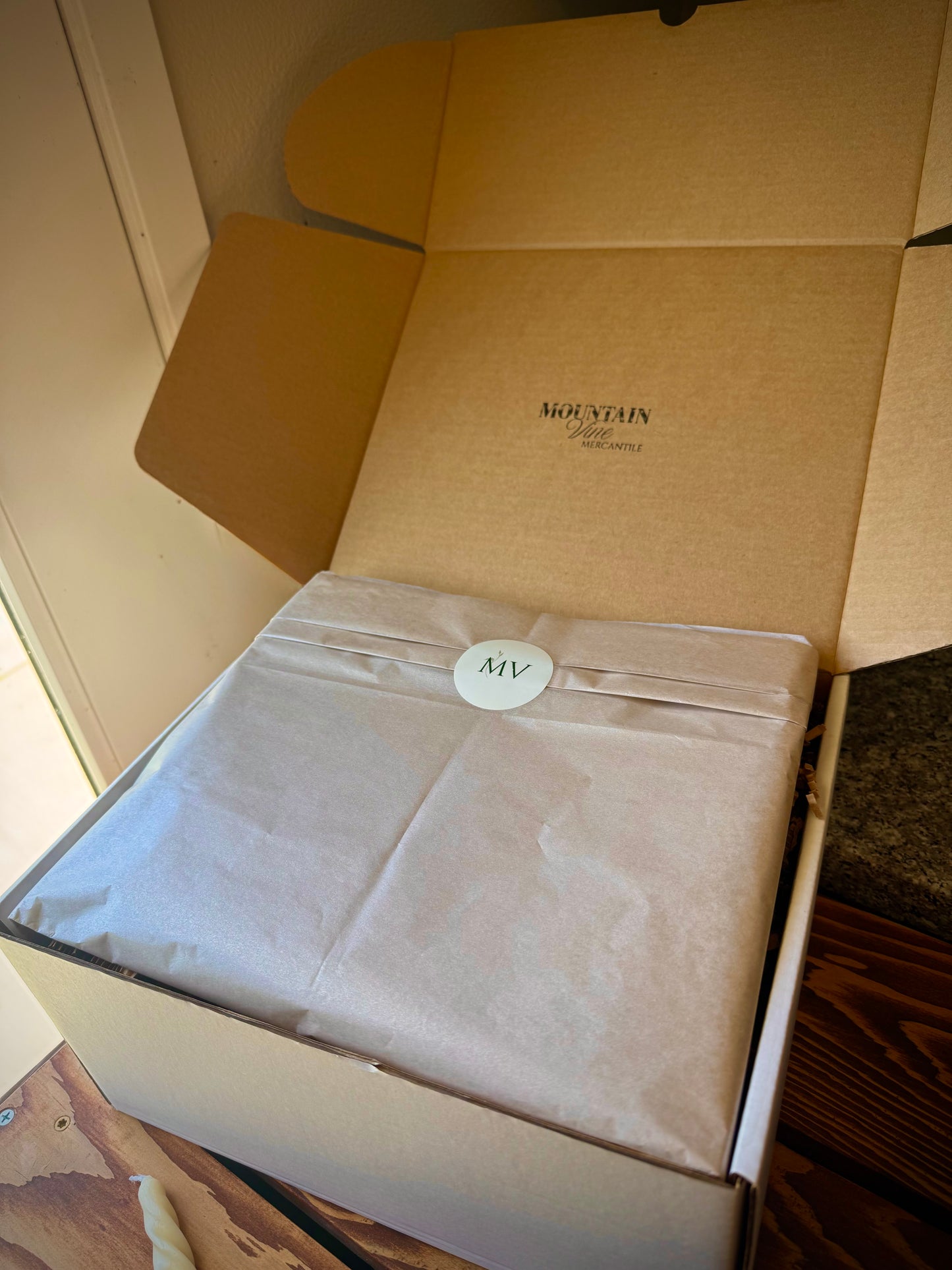 Mountain Vine Mercantile gift box displayed on a rustic wood table opened with inner logo showing and champagne colored wrapping tissue with MV logo sticker.
