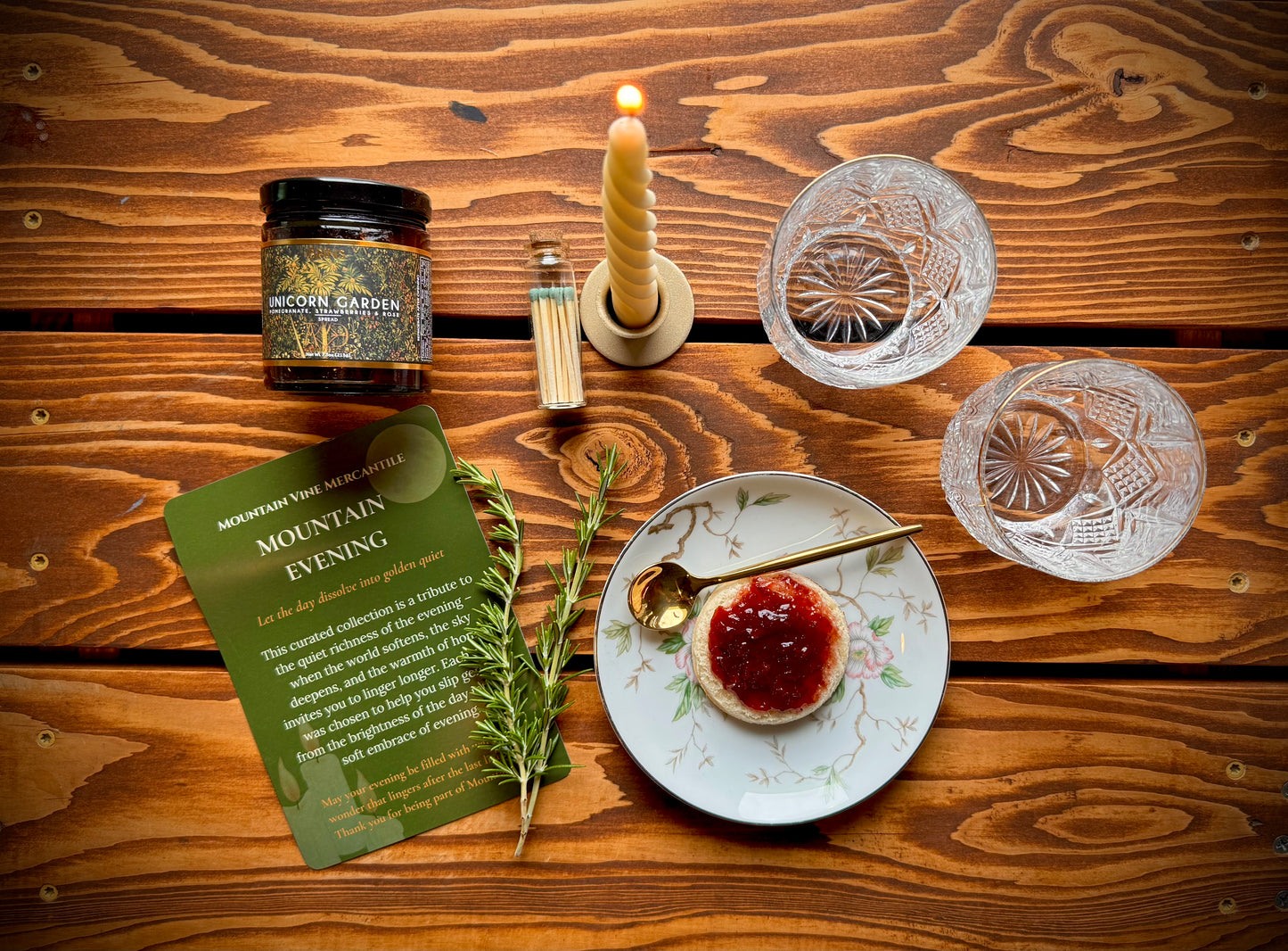 Mountain Vine Mercantile's Mountain Evening gift box display with jam topped biscuit, floral plate, candle rosemary spring, crystal glasses and product card on rustic wood table.