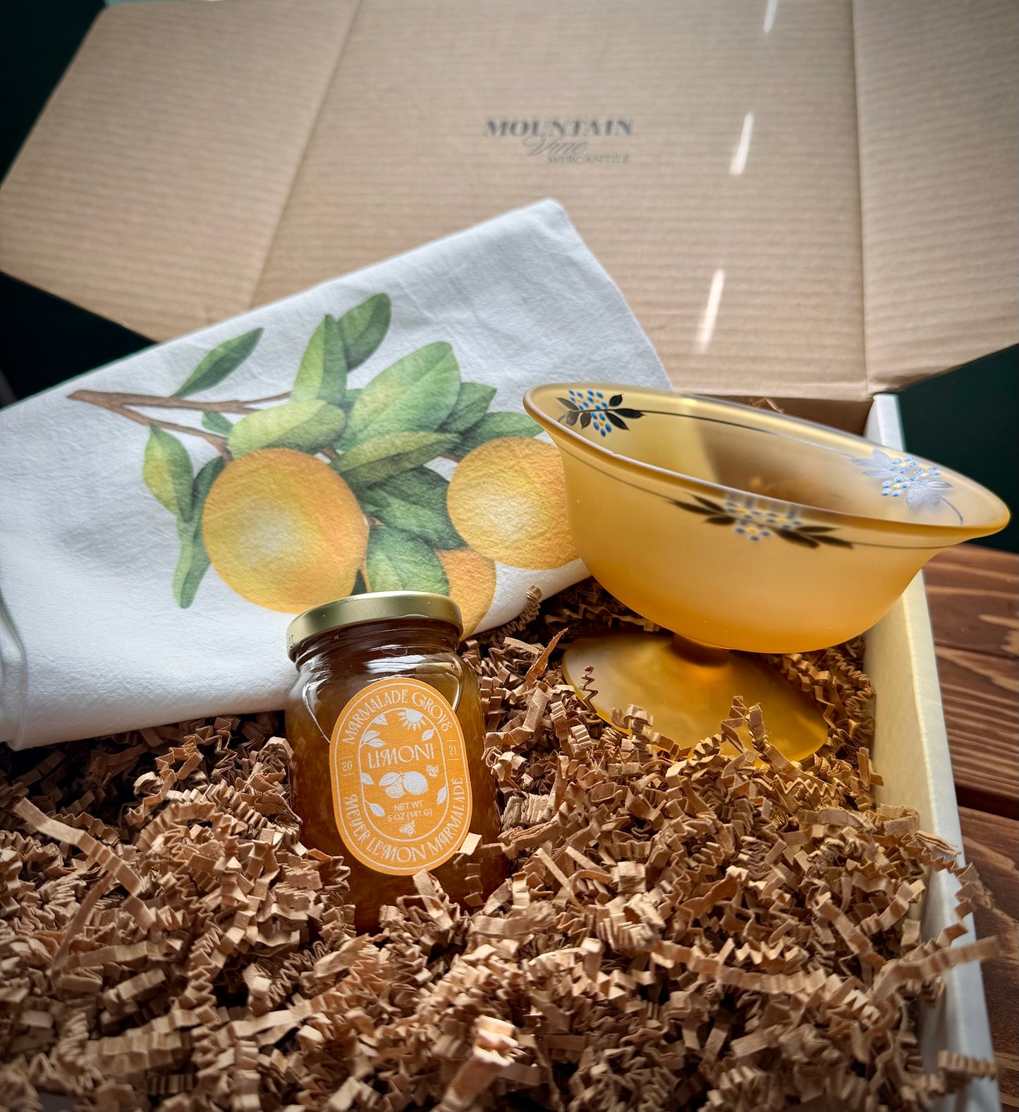 A jar of honey Meyer lemon marmalade, a lemon printed tea towel and a vintage frosted yellow compote on a bed of brown crinkle paper inside a Mountain Vine box