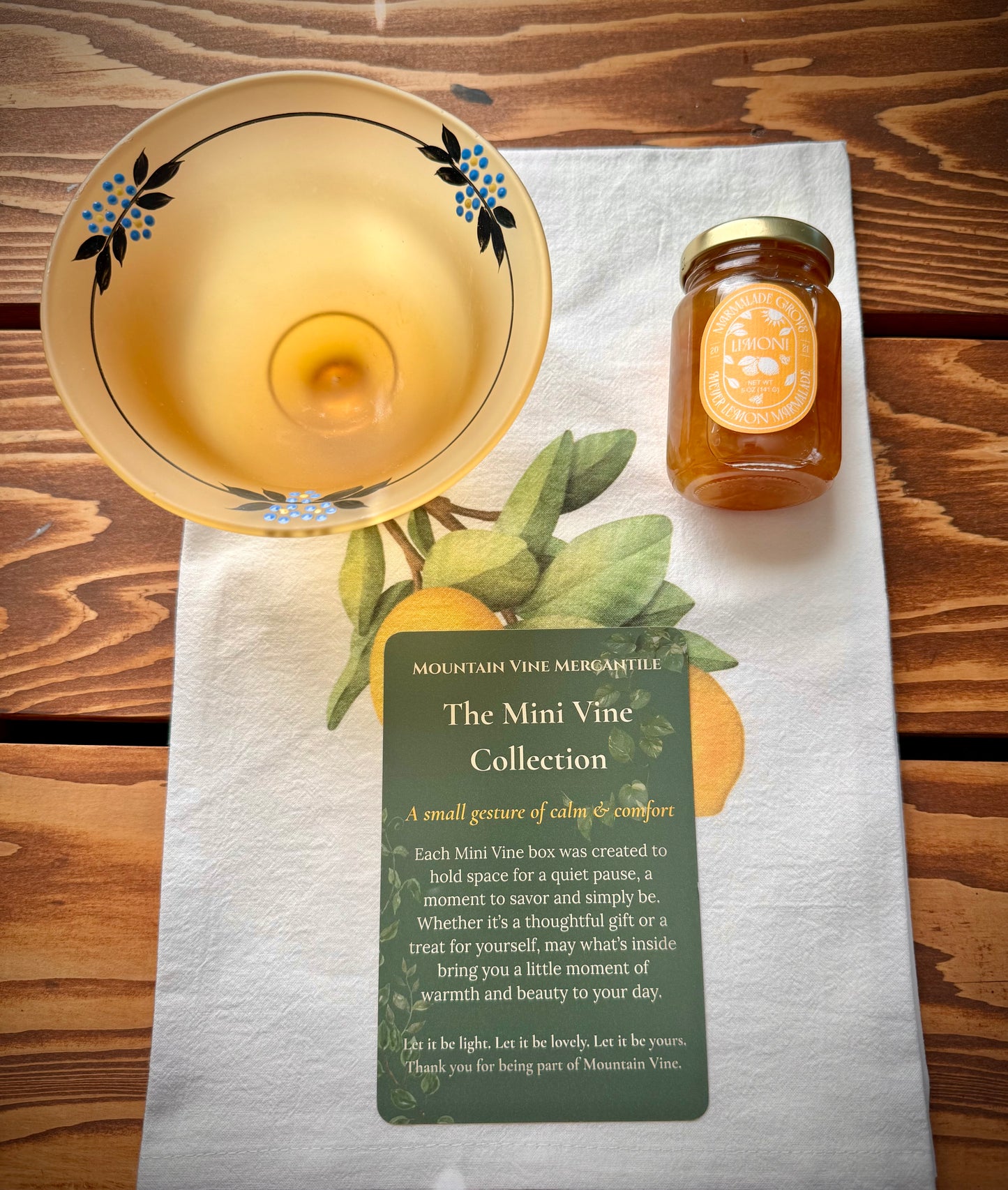 A vintage frosted yellow pedestal compote with painted blue flowers, a jar of honey Meyer Leon marmalade and a green Mini Vine collection insert card on top of a white tea towel on a wood surface