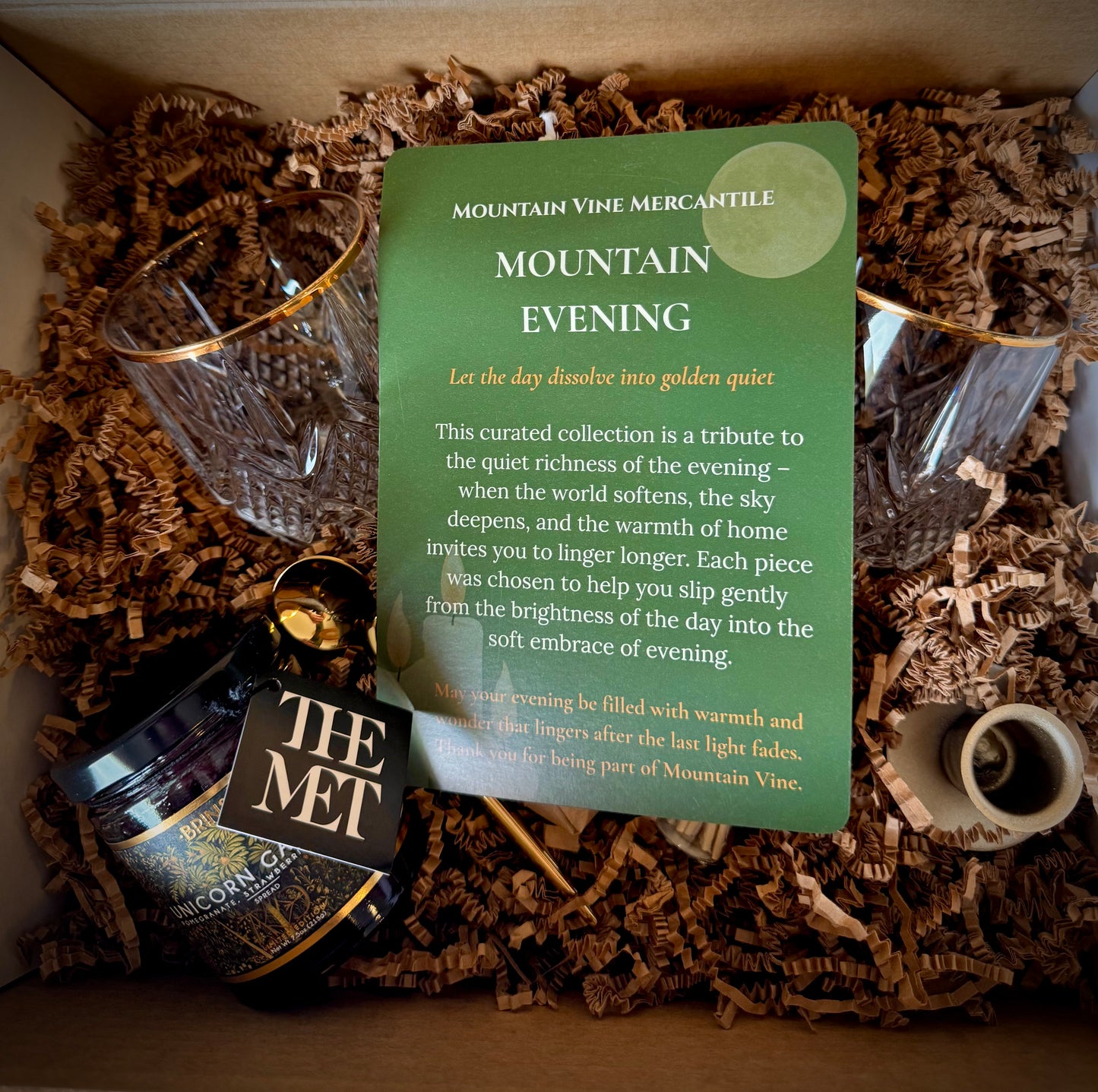 Mountain Evening gift box opened featuring the Mountain Evening insert card on a background of brown crinkle paper and box contents including vintage glasses, taper Holder and unicorn garden jam.