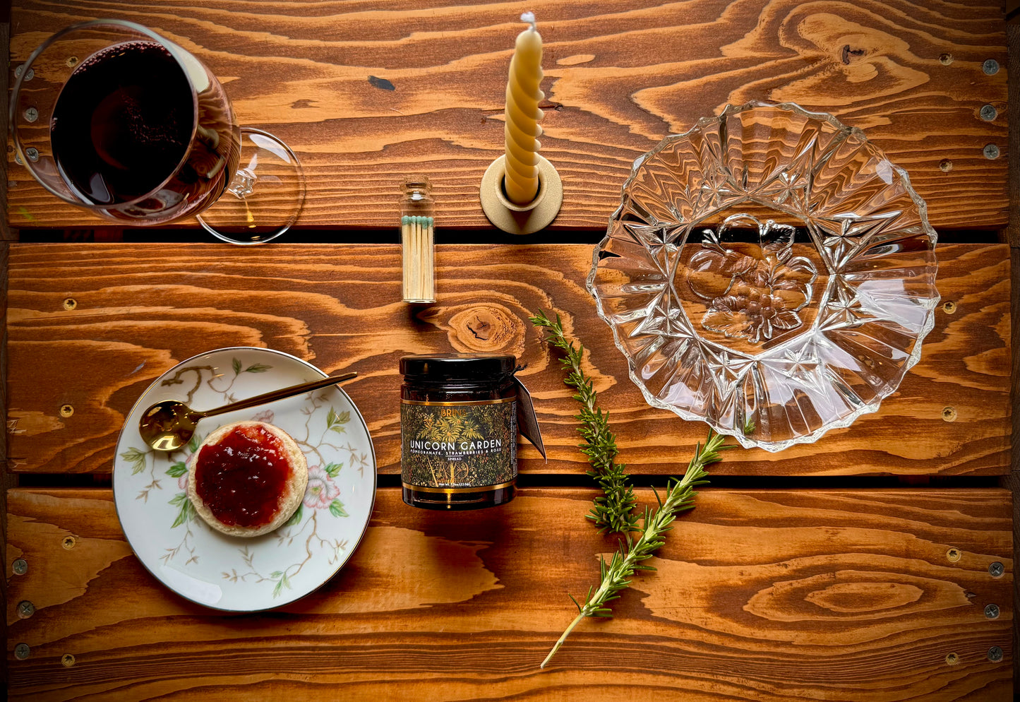 Mountain Vine Mercantile Mountain Evening gift box contents displayed on rustic wood table featuring a Unicorn Garden jam by BRINS, biscuit with jam on a floral plate, gold Spoon, glass of red wine, vintage clear scalloped edge glass bowl and lit candle in gold taper holder. 