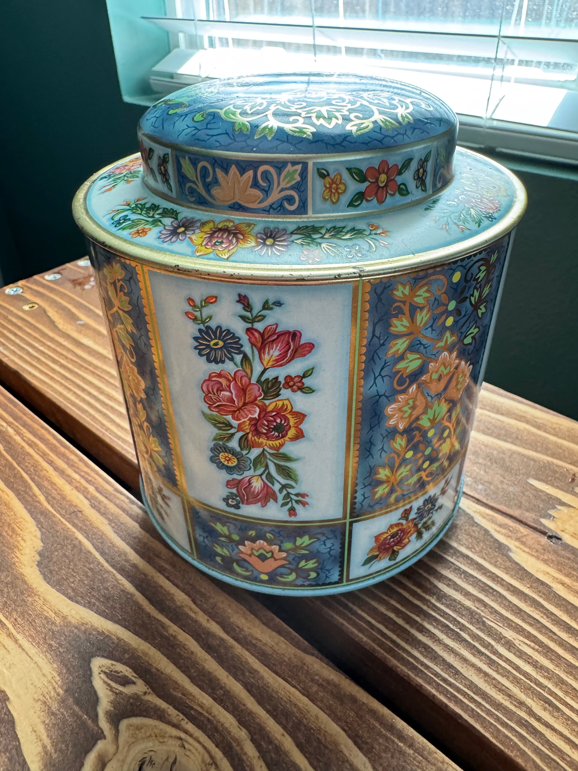 Close up angled view of a patchwork floral  english vintage tea tin on a wood table with a green wall and window in the background