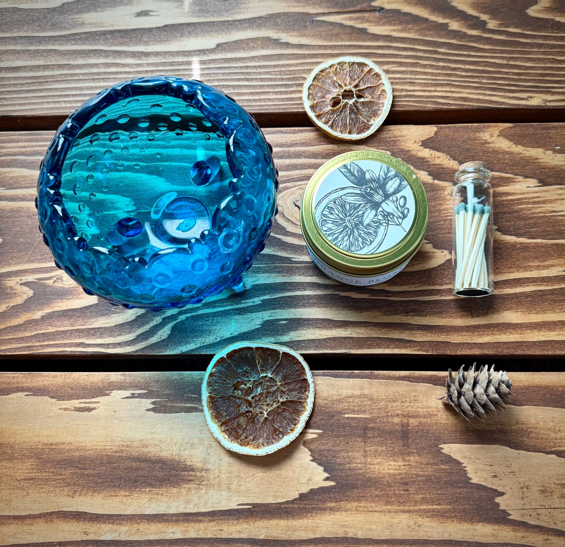 Flat lay of blue vintage hobnail glass bowl, botanical soy candle, match vial and dried citrus on a wood background. 