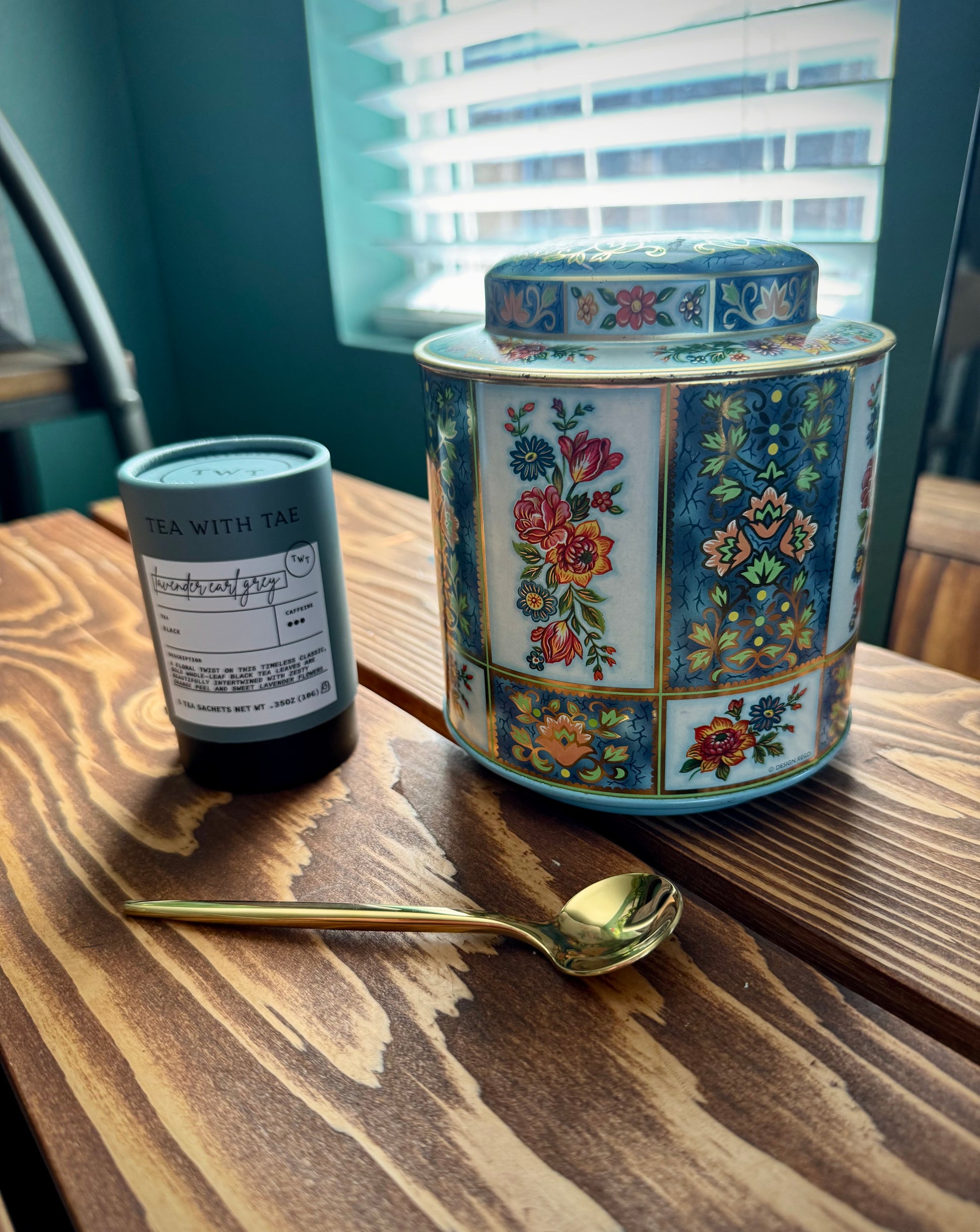 Vintage English floral tea tin by Daher next to lavender Earl grey tea and a gold spoon on a wood table in front of a window. 