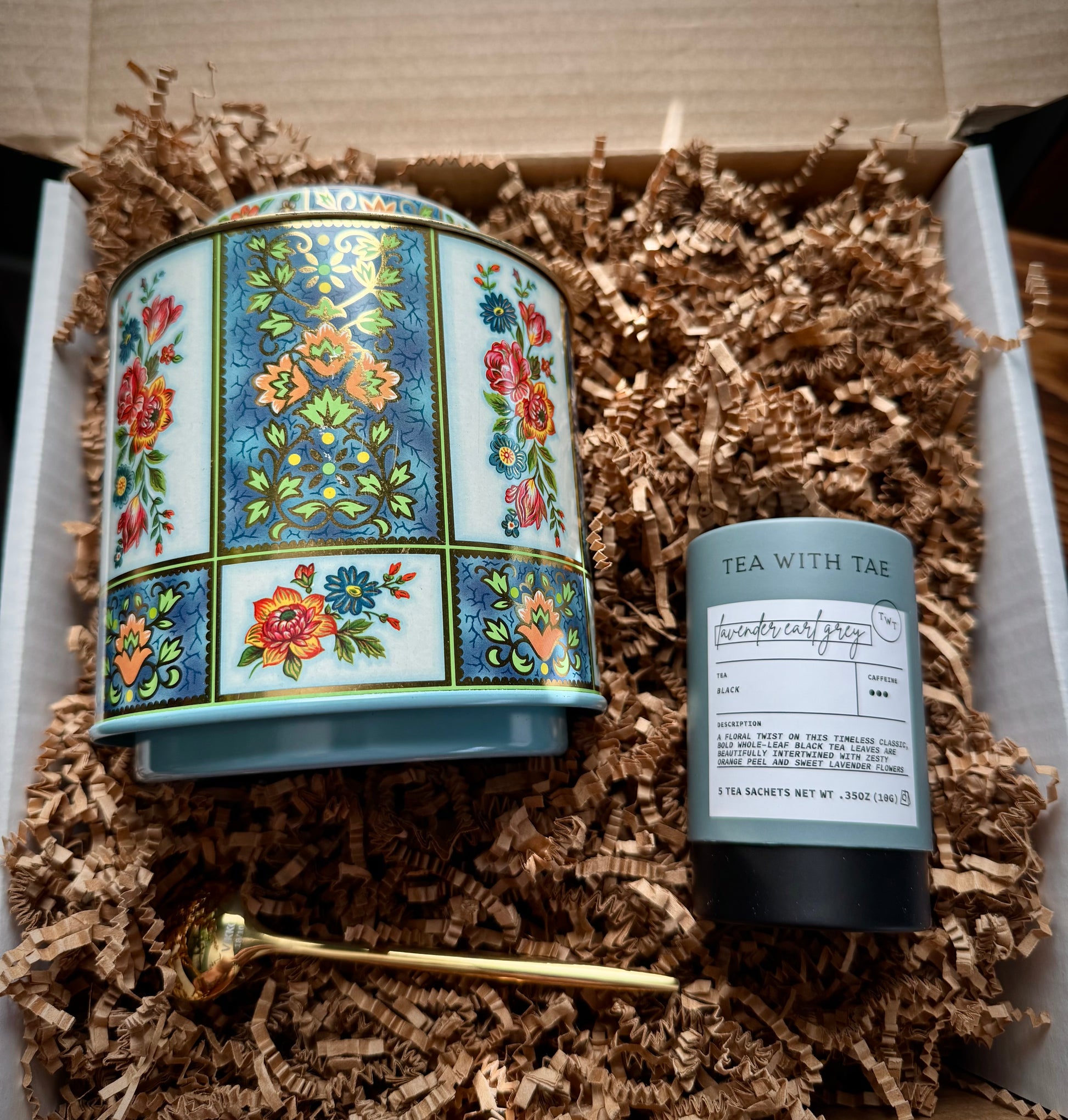 Blue, gold, green, orange and red floral tea tin, lavender Earl grey tea and a gold teaspoon on a bed of brown crinkle paper in a product box