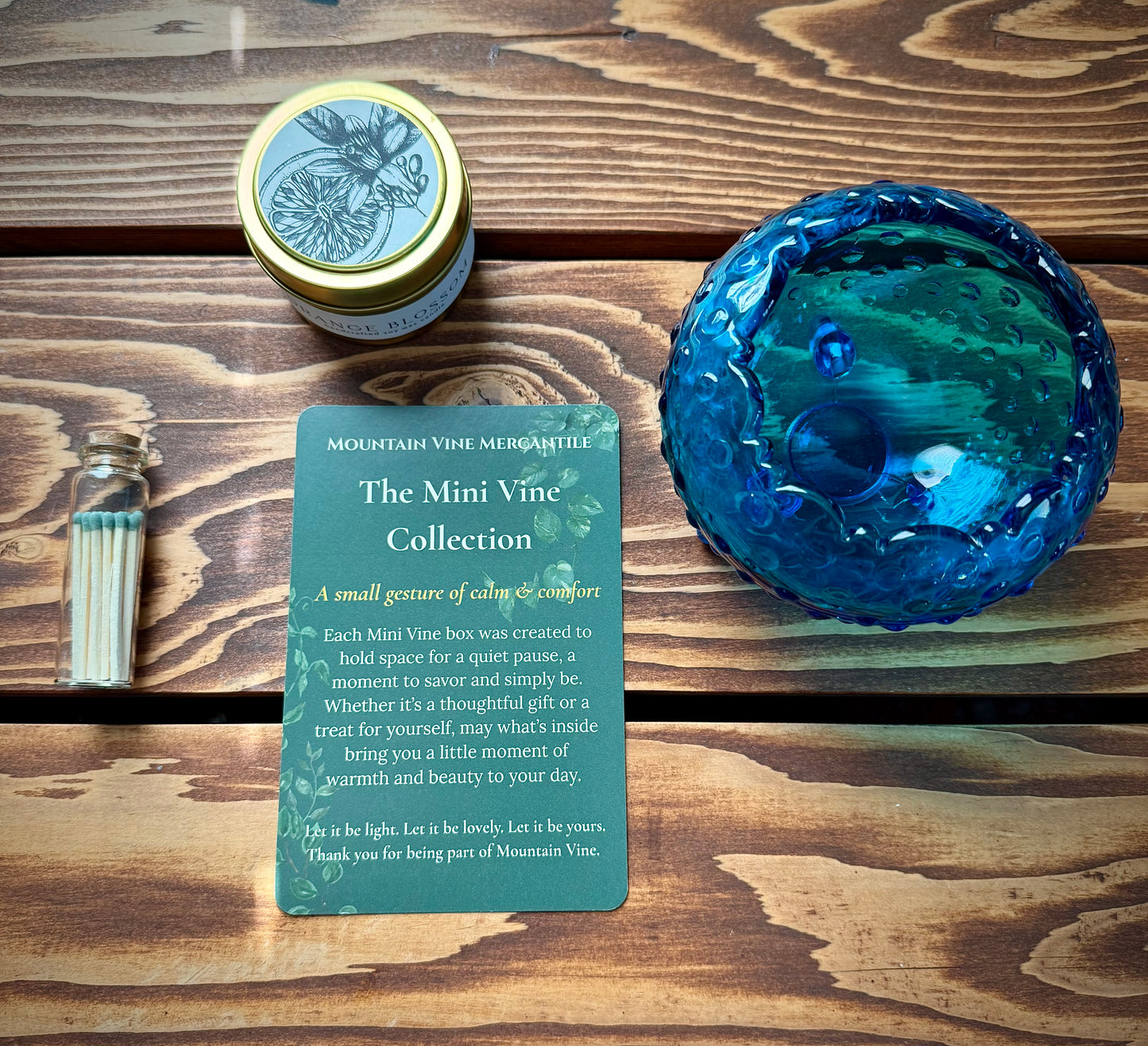 Green tipped match vial, gold tin soy candle, green Mii Vine Collection insert card and vintage blue hobnail dish on wood background.