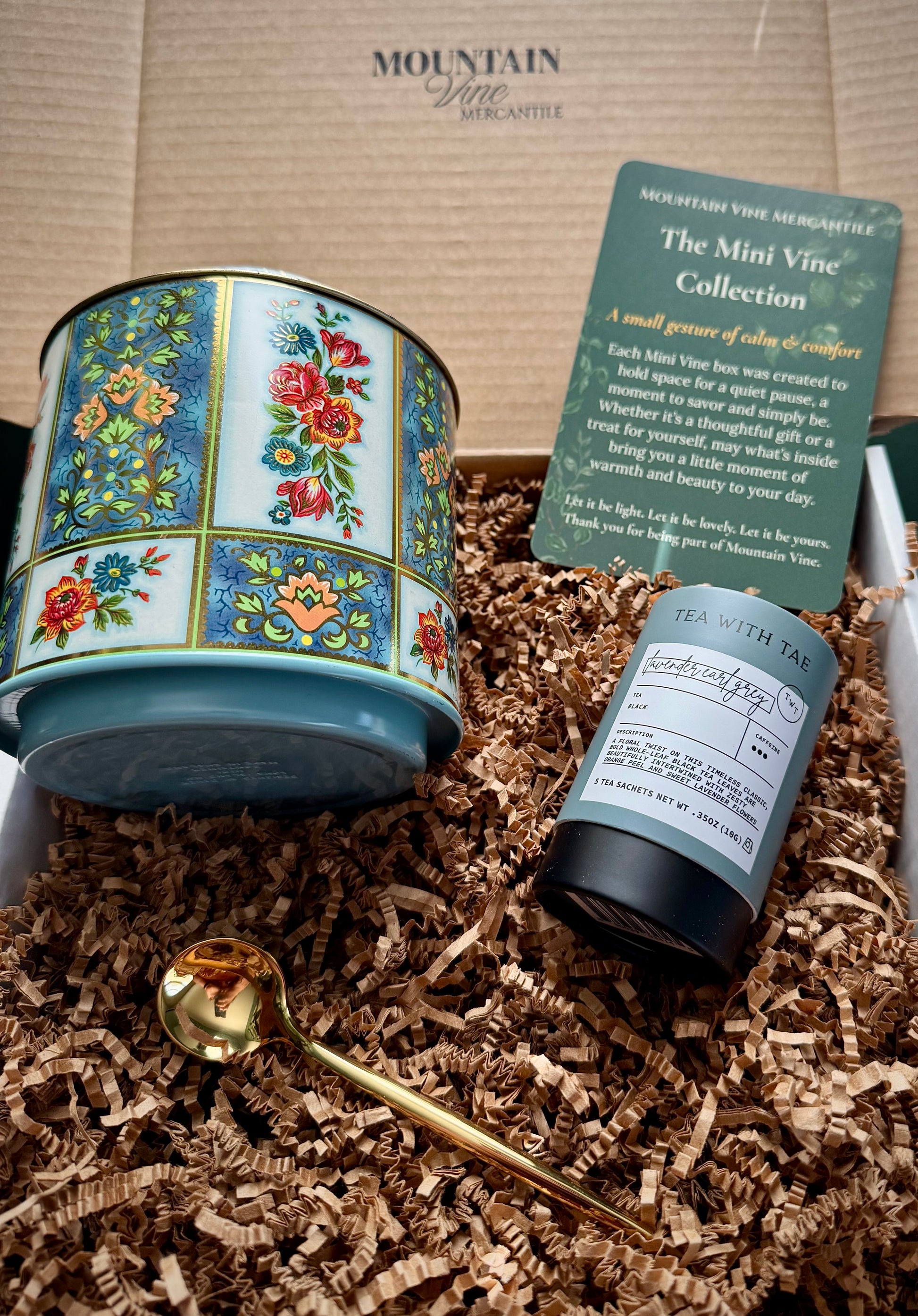 Blue floral English tea tin with lavender Earl grey tea from tea with tae, a shiny Gold teaspoon and a green product insert card on a bed on brown crinkle paper and inside a Mountain Vine Mercantile box
