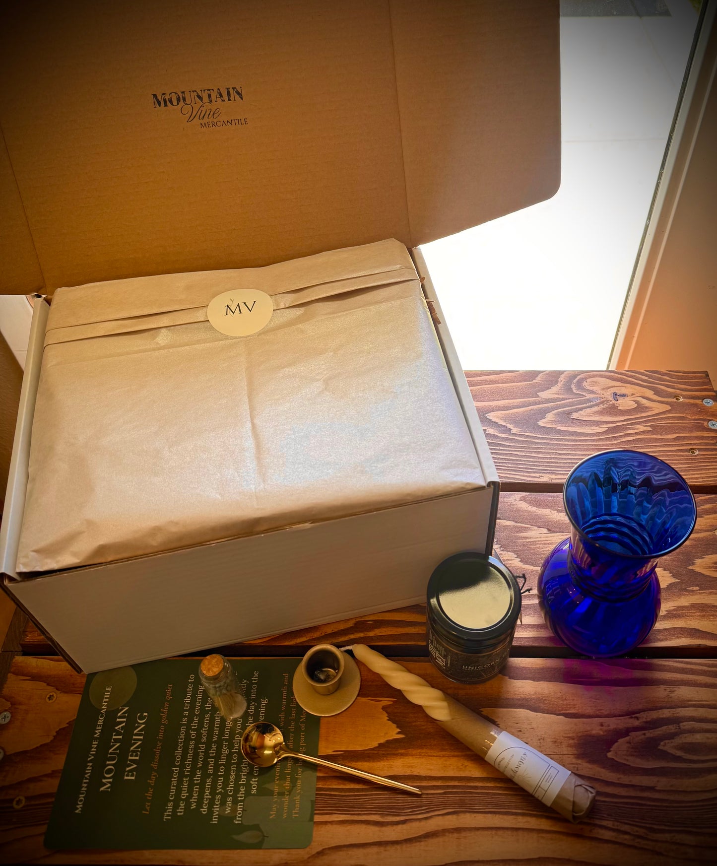 Mountain Evening gift box on table opened with champagne wrapping tissue and MV logo displayed. Contents of the box laid next to the box including insert card, gold Spoon, gold taper holder, match vial, unicorn Jam and a vintage blue glass vase on a rustic wood table. 