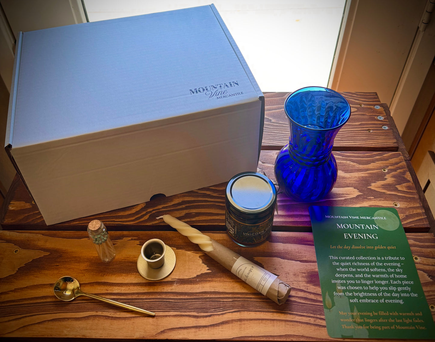 Mountain Vine Mercantile Mountain Evening closed gift box on a rustic wood table. The contents of the box are displayed next to it including the insert card, twisted candle, gold taper holder, gold spoon, match vial, unicorn garden jam and vintage blue vase