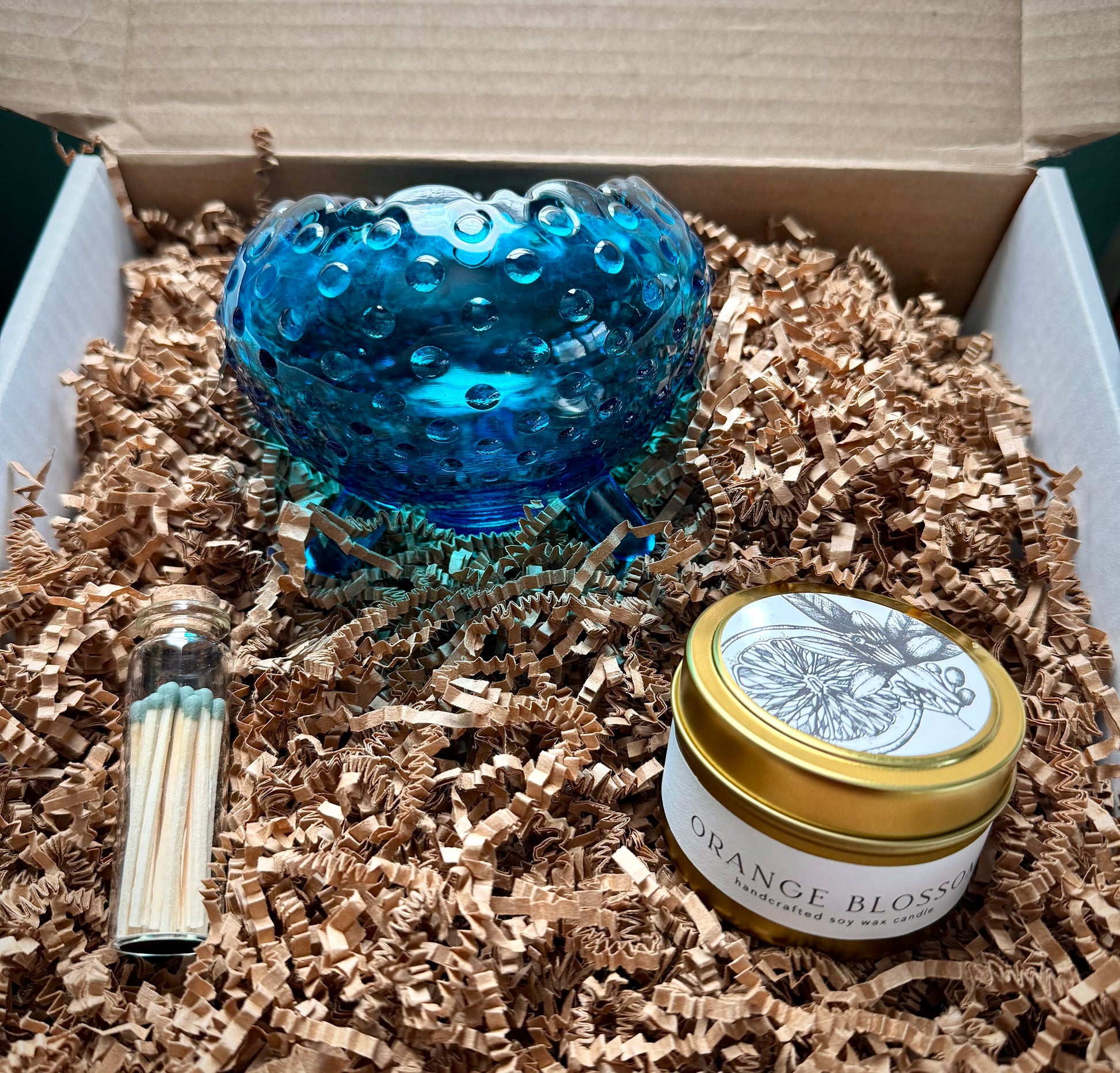 Vintage blue hobnail glass bowl, botanical soy candle and green tipped match vial on a bed of crinkle paper inside product box.