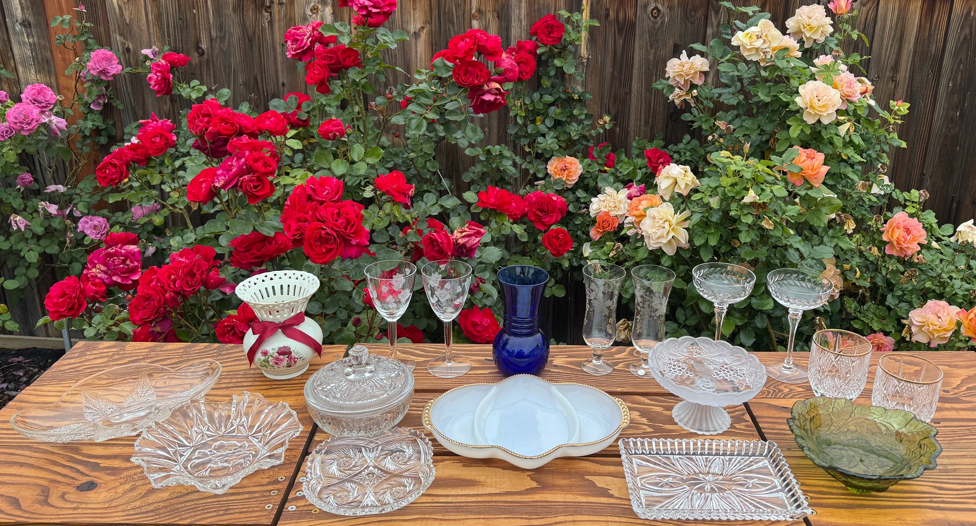 Assortment of vintage items for the Mountain Evening gift box by Mountain Vine Mercantile. The assortment includes a mix of glass and ceramic bowls, lidded dishes, glassware and vases in shades of clear, white, blue, green and red on a rustic wood table with roses in the background. 