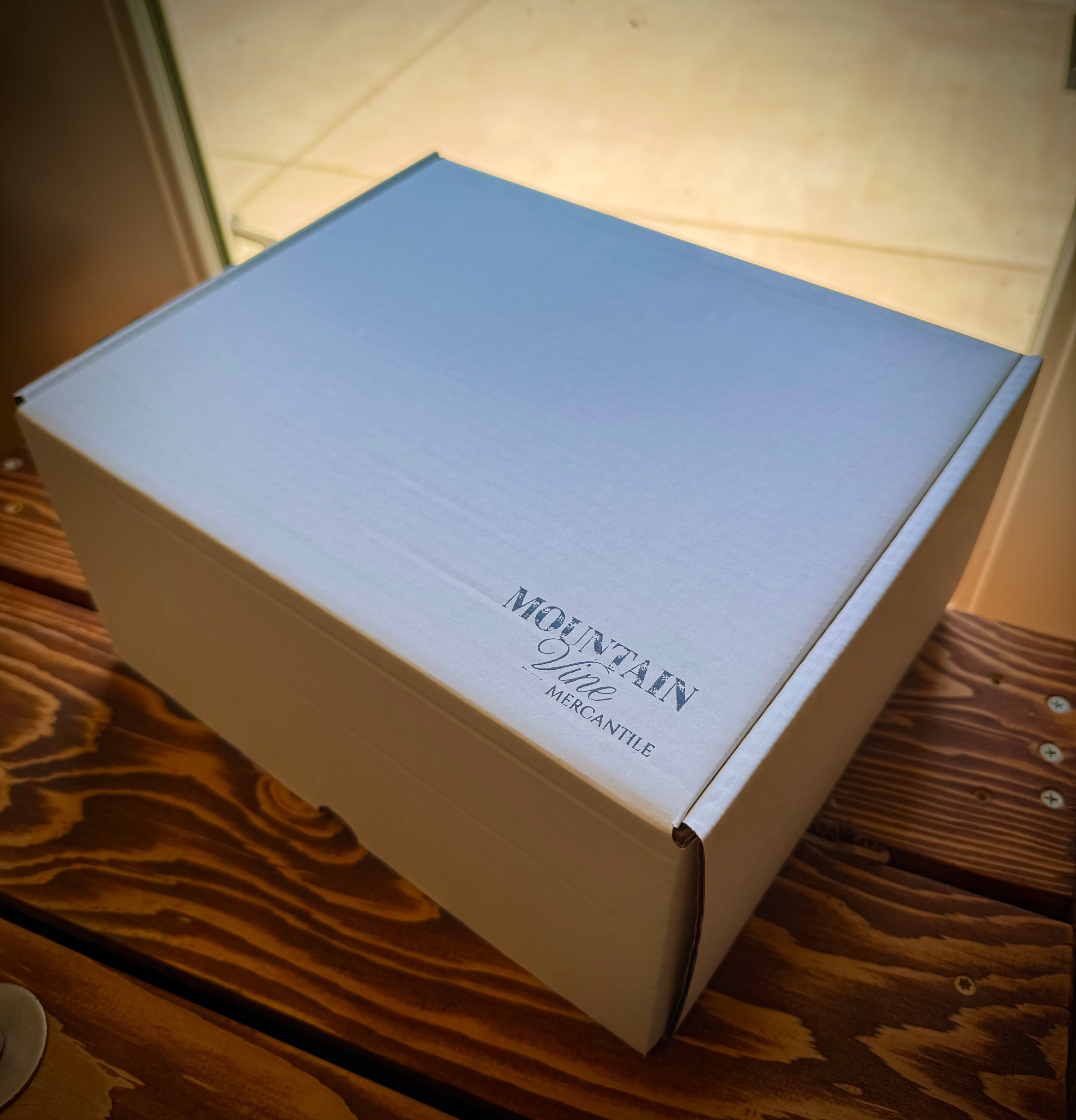 Mountain Vine Mercantile closed white gift box with the Mountain Vine Logo in the corner. Box is resting on a rustic wood table