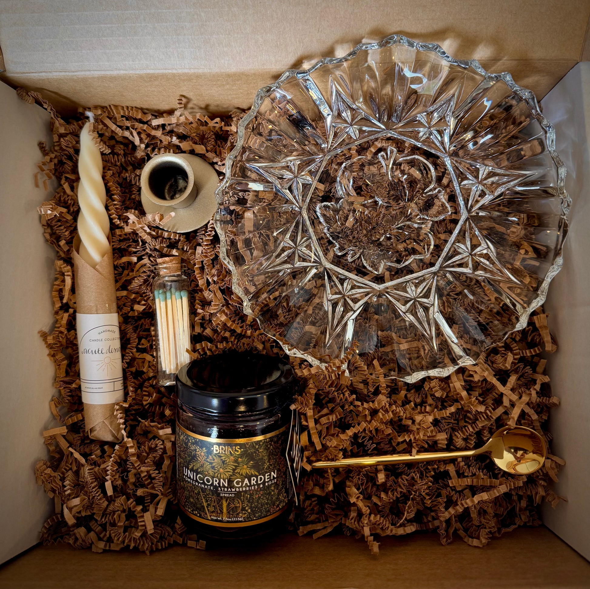 Mountain Vine Mercantile Mountain Evening box open showing contents of crinkle paper with twisted taper candle, green tippped matches in a vial, unicorn garden jam, gold sppon, taper Holder and clear glass scalloped edge bowl.