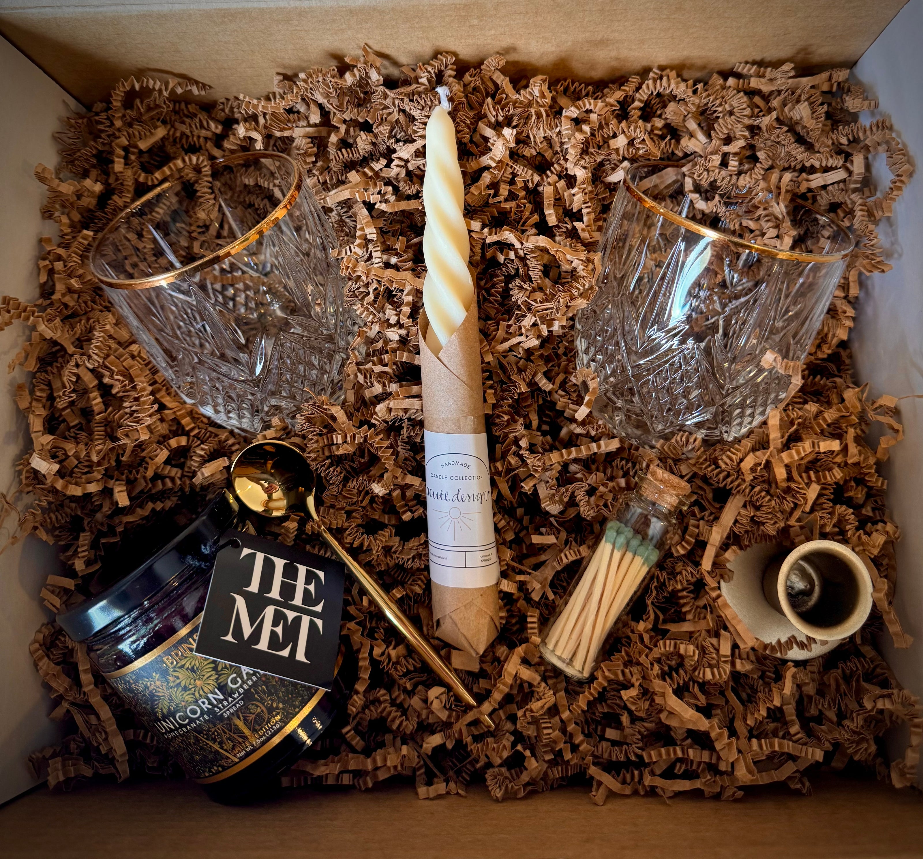 Mountain Vine Mercantile Mountain Evening open gift box showing packing crinkle paper and gift contents: vintage crystal glasses, hand twisted taper candle, gold taper Holder, match vial with green tippped matches, unicorn garden jam, and gold sppon