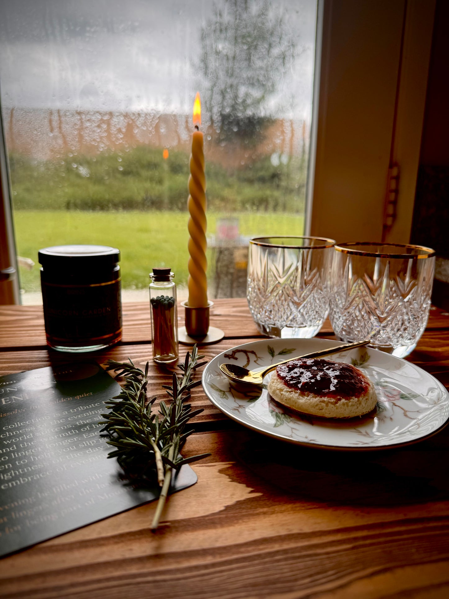 Mountain Evening gift box contents in use.  Two crystal glasses on a rustic wooden table with a lit hand twisted candle in a gold taper holder, spring of rosemary, unicorn Jam, and biscuit with jam on a floral plate in the foreground. Mist on window in background.
