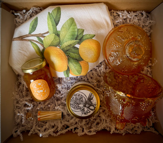 Mountain Vine Mercantile Mountain Day box open with an orange carnival glass vintage lidded candy dish, orange blossom candle in gold tin, limoni marmalade, match vial and a lemon branch tea towel on a bed of white crinkle paper