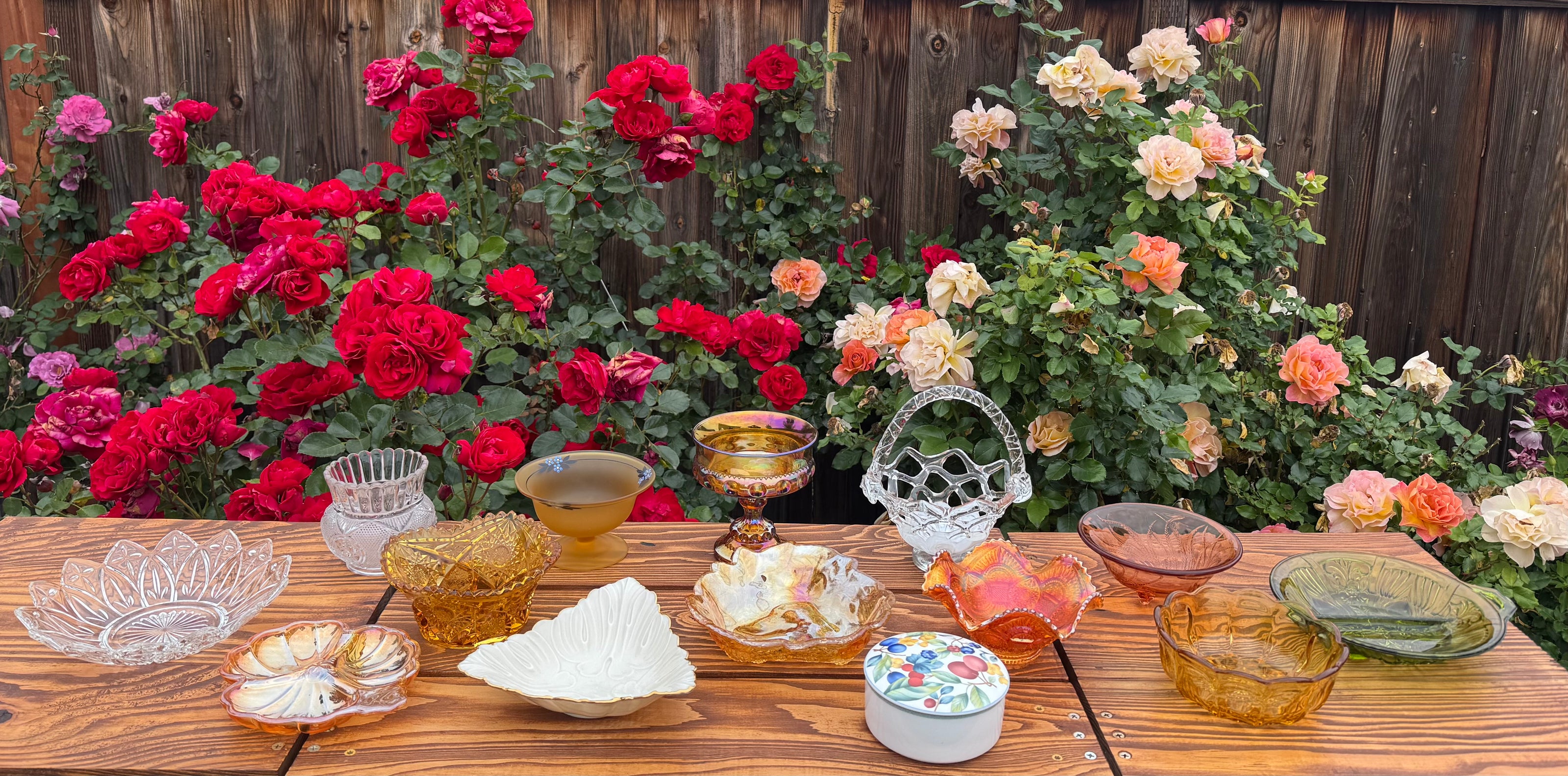 Mountain Day Gift box assortment of vintage glass and ceramic bowls, baskets vases, plates in hues of orange, gold, green, clear and white with a rose background on a rustic wood table