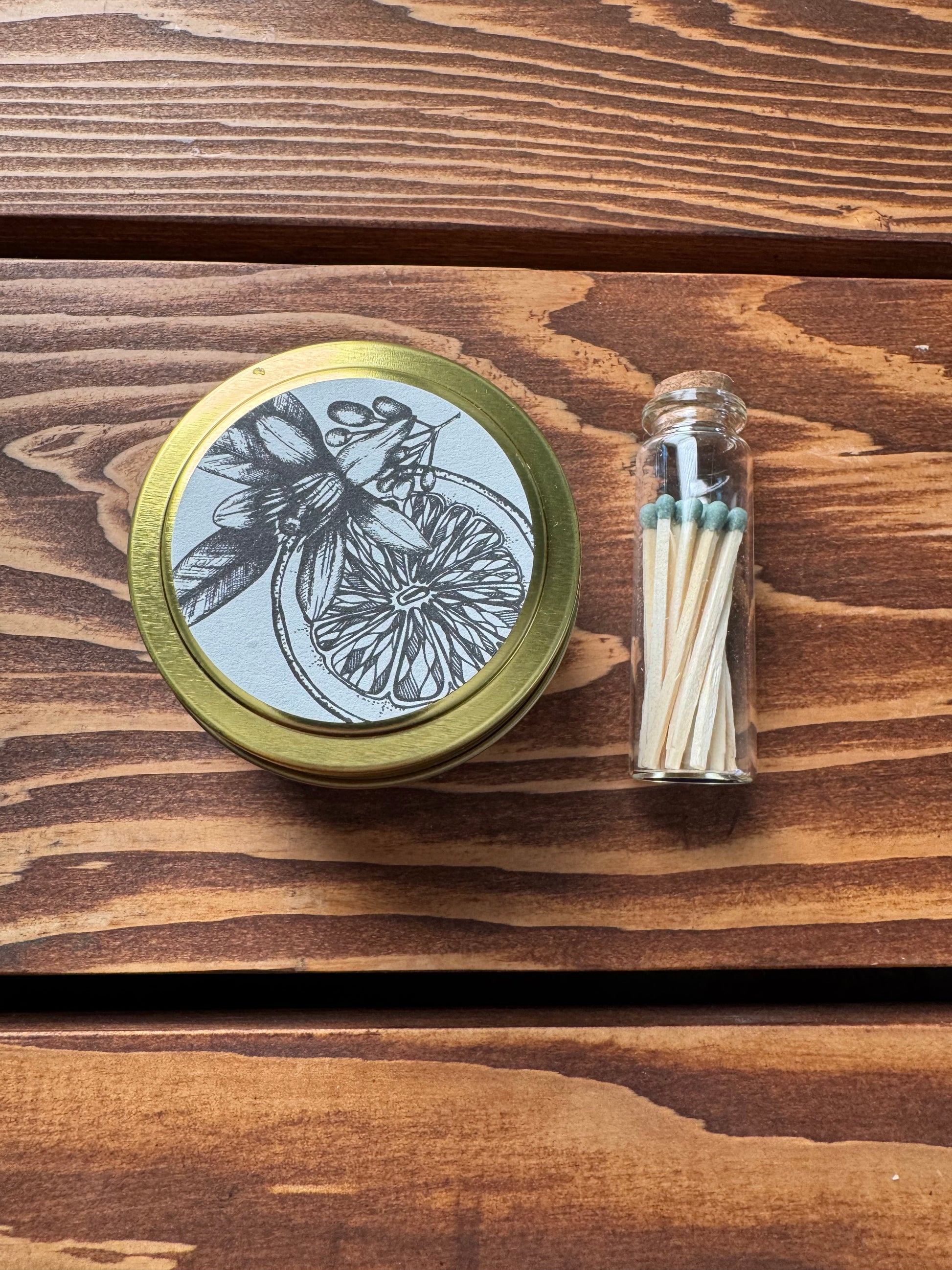 Close up shot of an orange blossom soy candle and green tipped match vial on a wood background. 