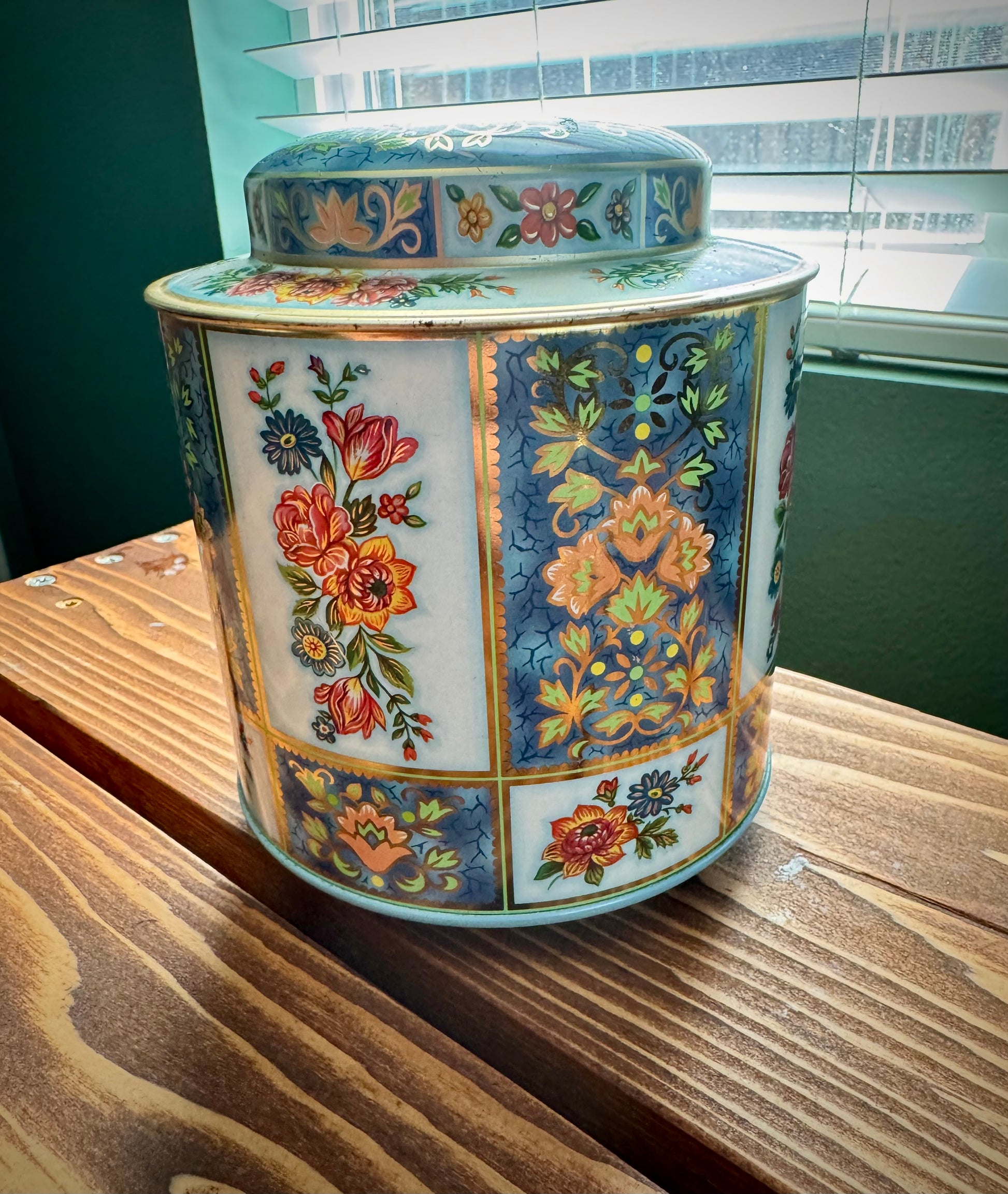 Close up view of vintage blue and gold floral patchwork tea tin by Daher on a wood surface with window in the background