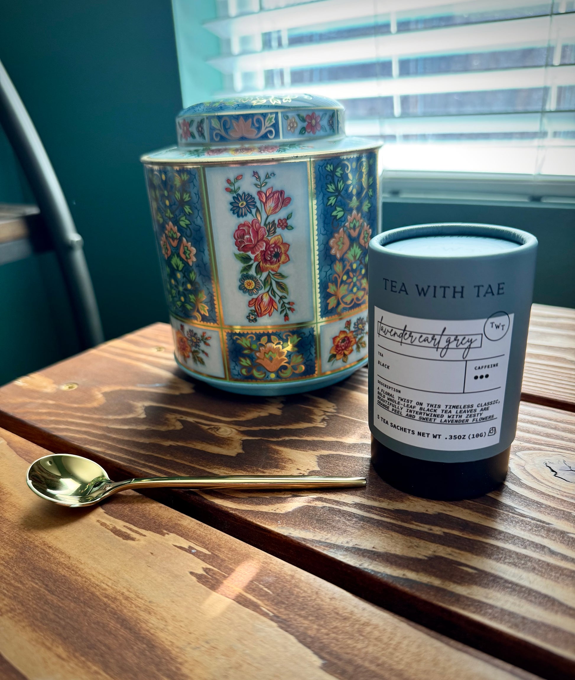 Angled shot of a vintage blue floral English tea tin, Lavender Earl Grey Tea in blue cylindrical packaging and a gold teapoon on a wood surface in front of window with blinds. 